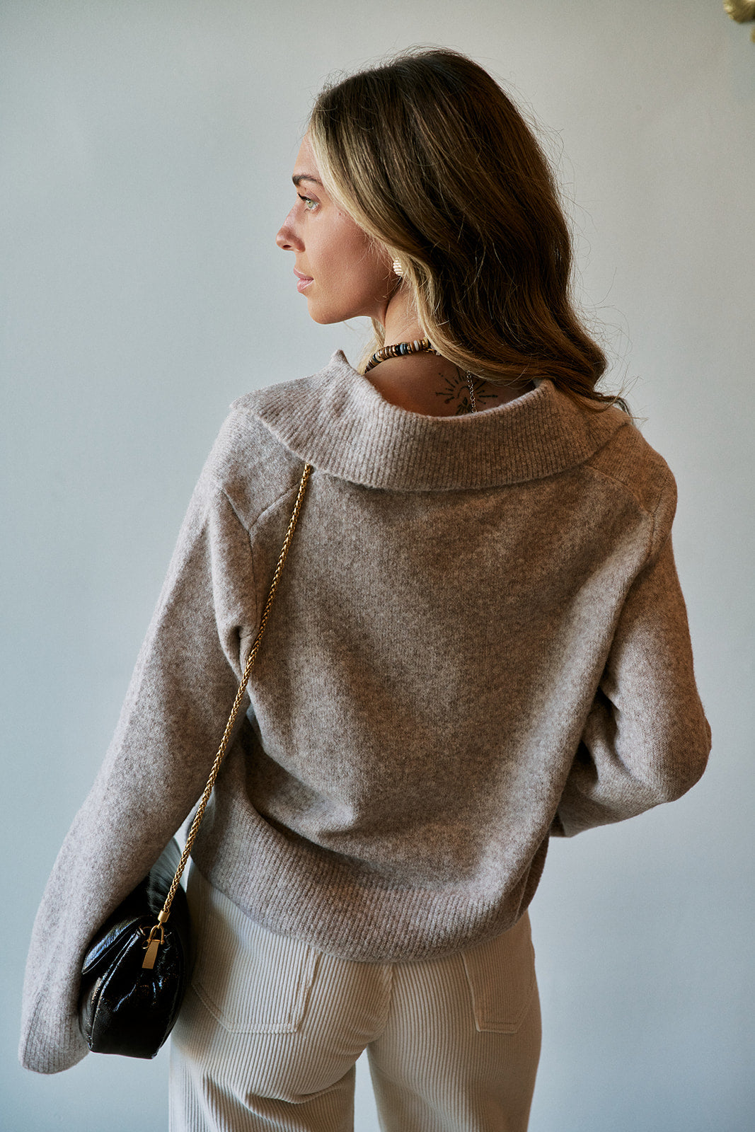 A woman wearing a light taupe knit sweater and cream pants, shown from a side-back angle against a plain light gray wall. She is carrying a small black shoulder bag with a gold chain strap.
