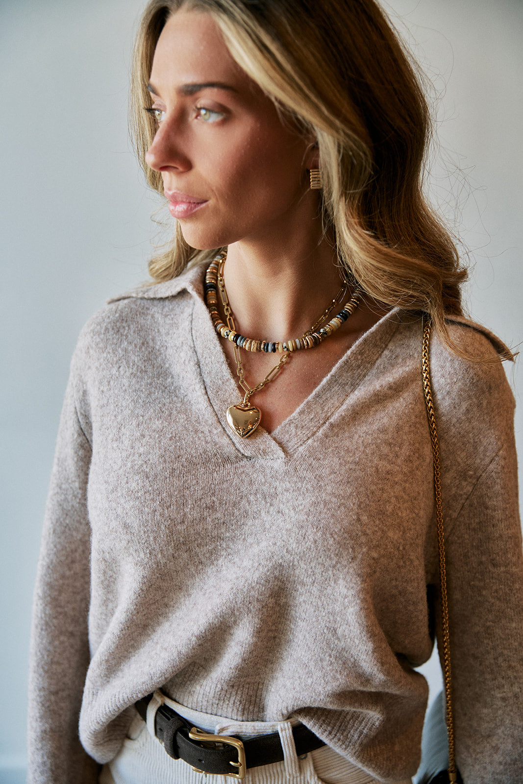 A woman wearing a light beige knit V-neck sweater, layered gold necklaces, and a black belt, shown in a close-up lifestyle shot against a neutral light gray background.