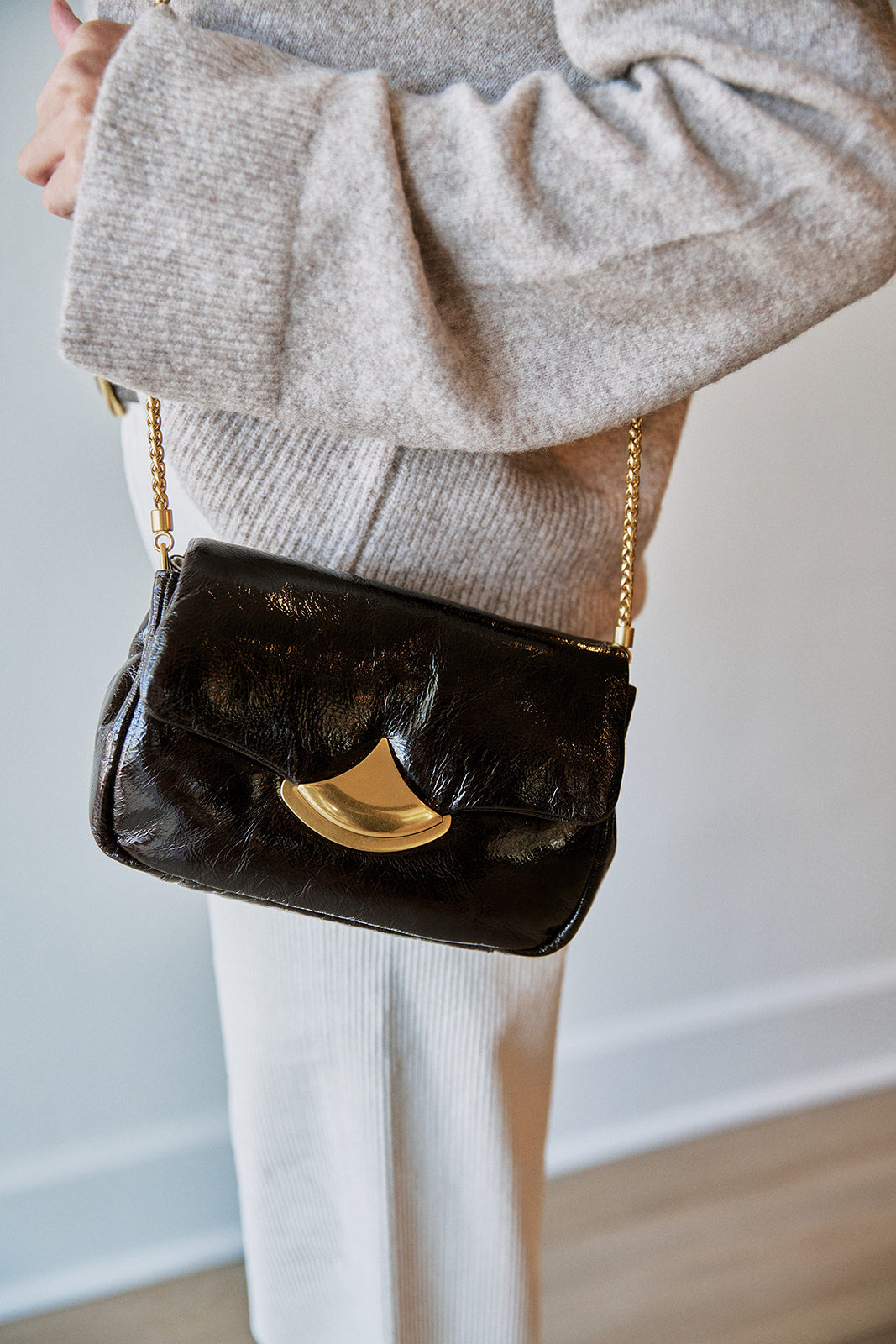 A person wearing a light beige knit sweater and white pants carries a dark textured leather shoulder bag with a gold crescent-shaped front detail and a gold chain strap.