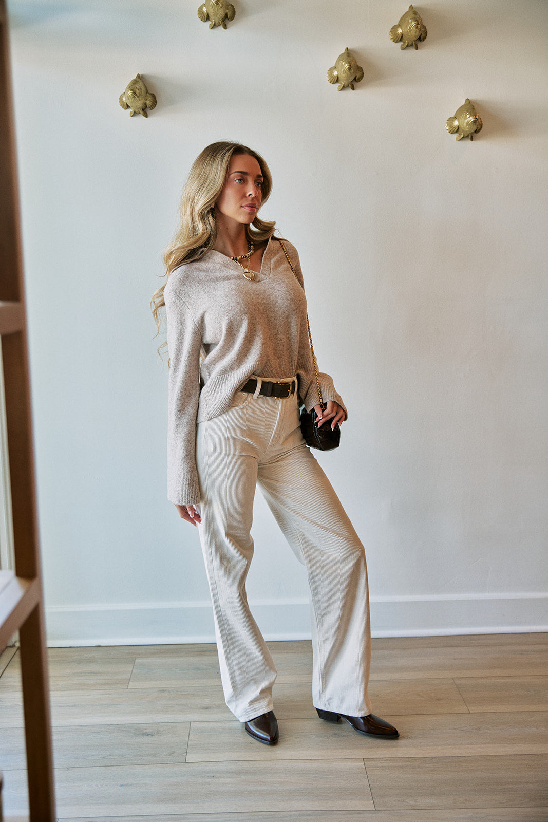 A woman modeling a cozy beige knit long-sleeve top and cream wide-leg trousers, styled with a black belt, black shoes, and a small black clutch, standing in a room with light wood floors and a white wall decorated with gold wall art.
