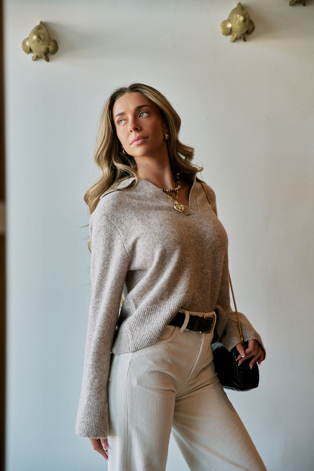 A woman wearing a light beige knit sweater and cream trousers, holding a small black handbag, standing against a white wall with decorative gold wall fixtures.