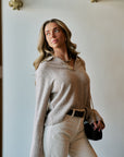 A woman wearing a light beige knit sweater and cream trousers, holding a small black handbag, standing against a white wall with decorative gold wall fixtures.