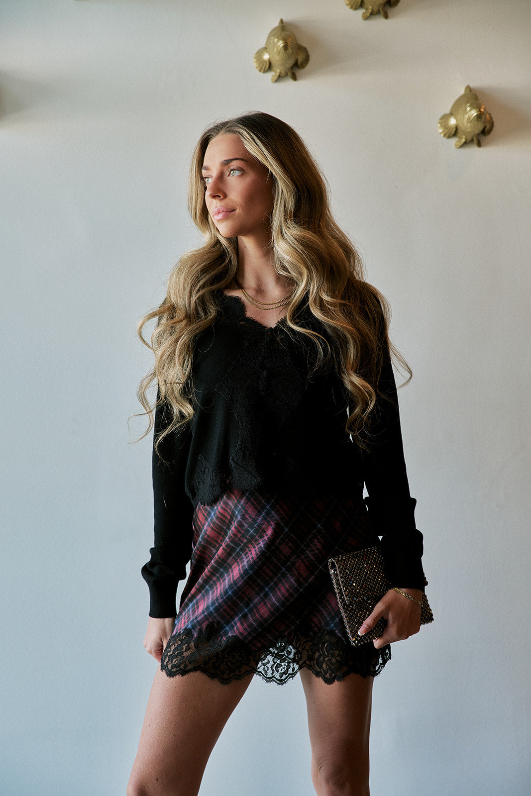 A woman stands in a room wearing a black long-sleeve ruffled blouse, a red and blue plaid mini skirt with black lace trim, and holding a small clutch.