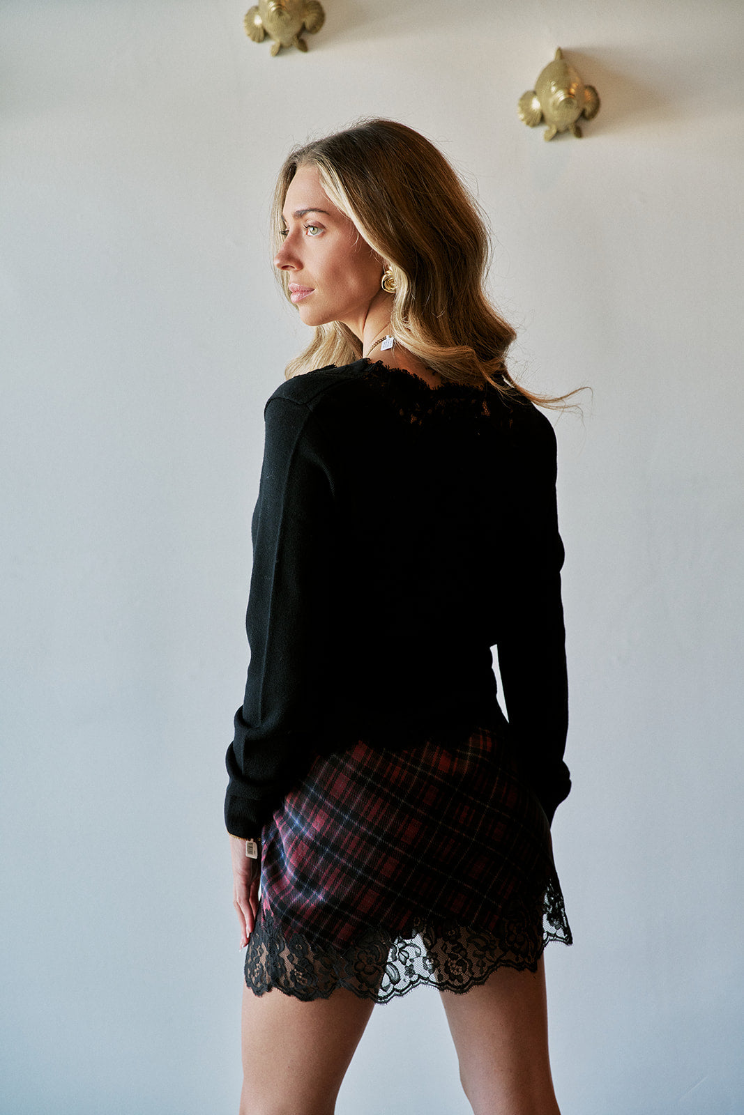A woman wearing a black long-sleeve knit top and a red and black plaid mini skirt with a black lace hem, photographed from the back against a light gray wall.