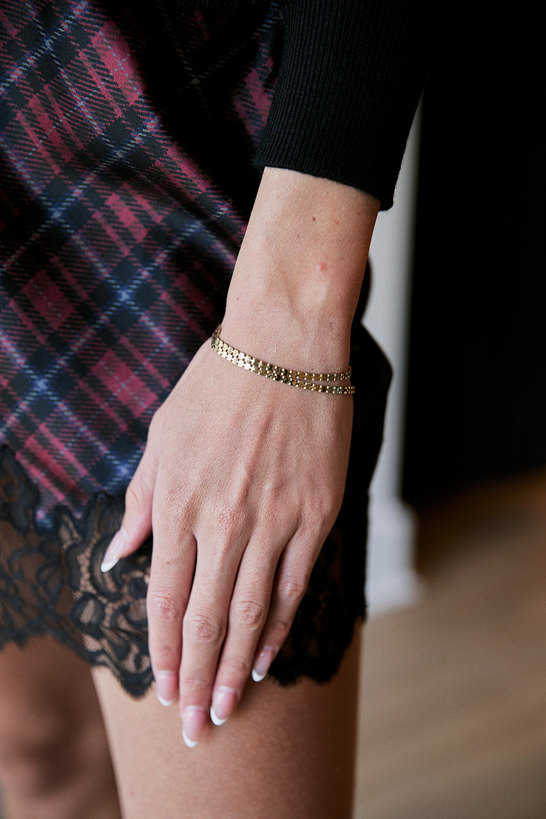 A close-up of a person's hand and wrist wearing a delicate double gold chain bracelet, styled with a black long-sleeve top, a red and blue plaid skirt, and a black lace hem against a dark background.