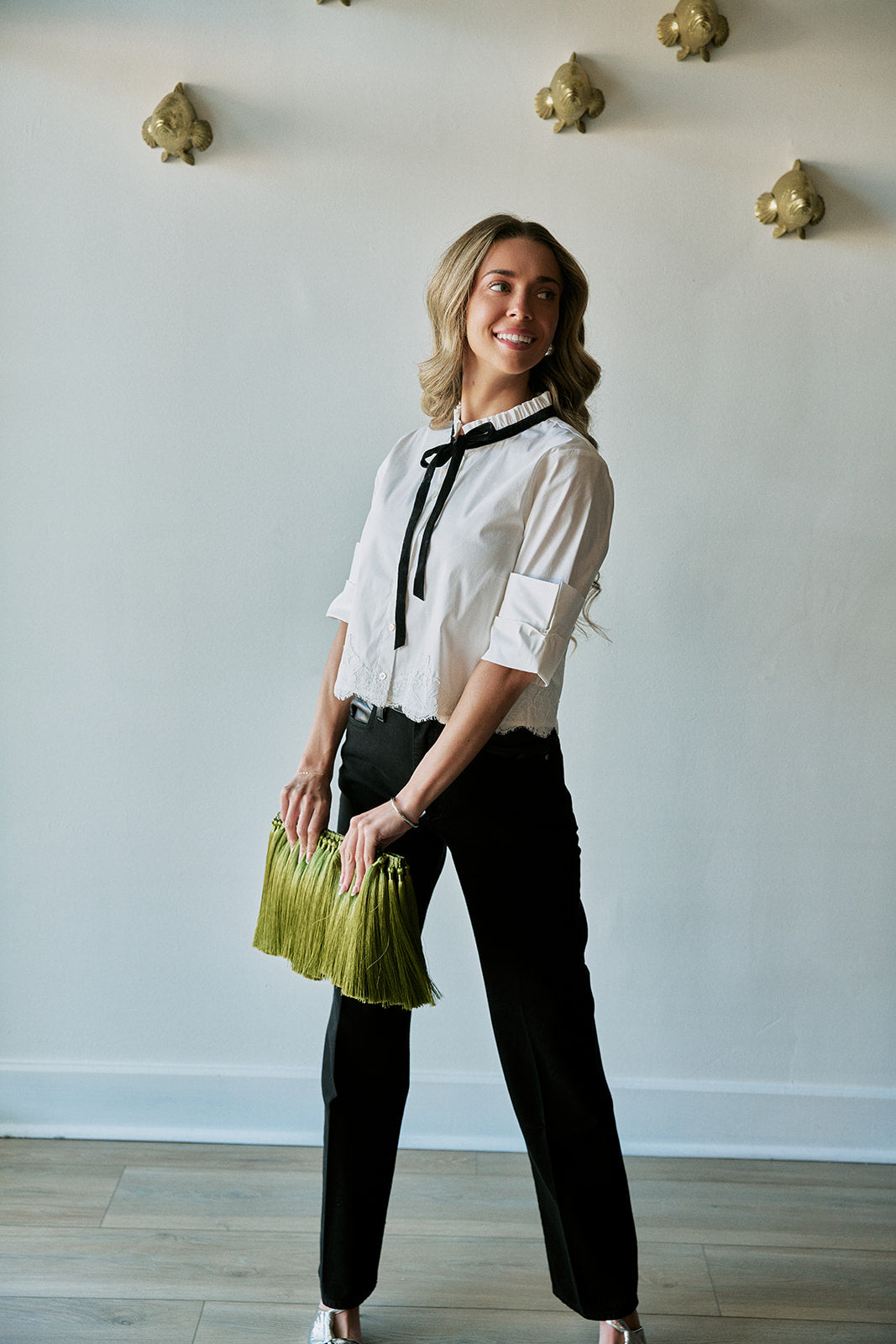 A woman modeling a white blouse with a black bow tie collar, paired with black pants and holding a green fringe clutch, standing against a white wall with gold decorative accents.