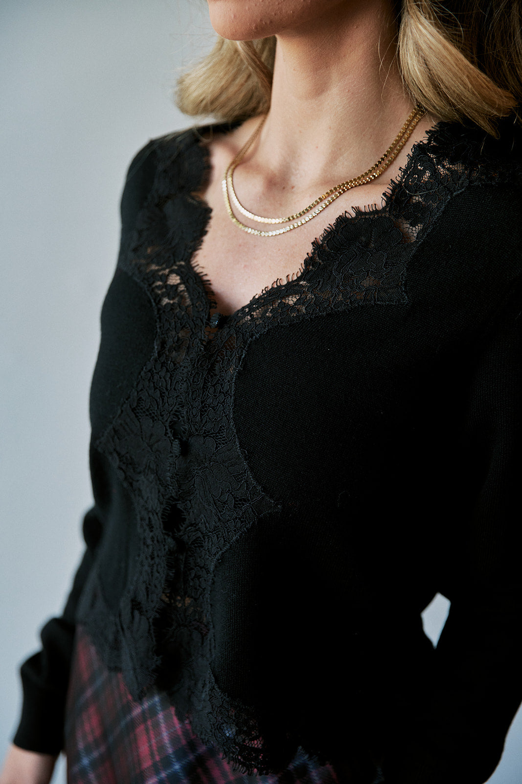 A close-up of a model wearing a black knit top with intricate black lace appliques and scalloped edges, styled with a gold chain necklace and a red plaid skirt against a neutral gray background.