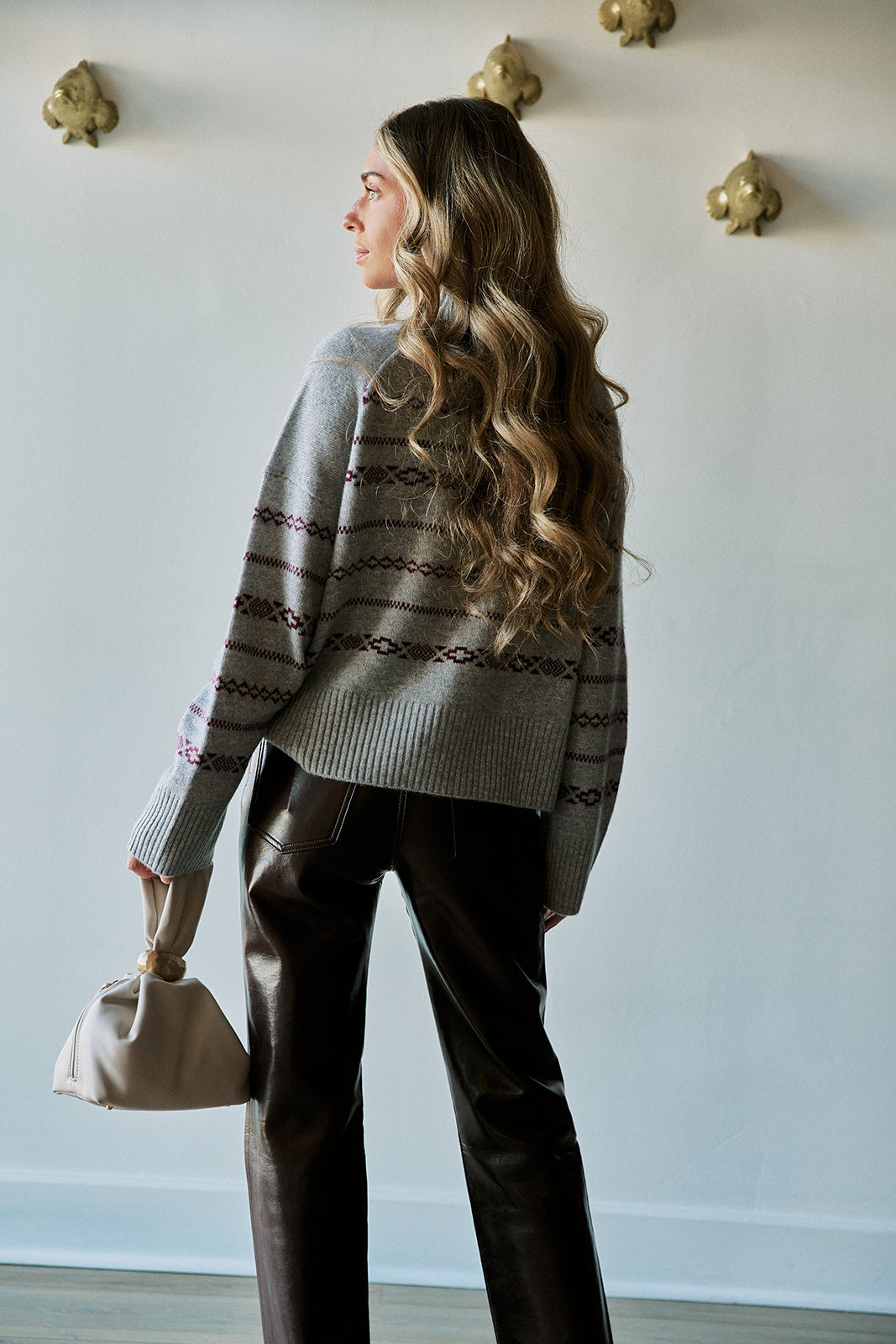 A woman with long wavy brown hair is shown from the back wearing a gray knit sweater with dark brown and burgundy geometric patterns, paired with dark brown leather pants and holding a beige handbag against a white wall.