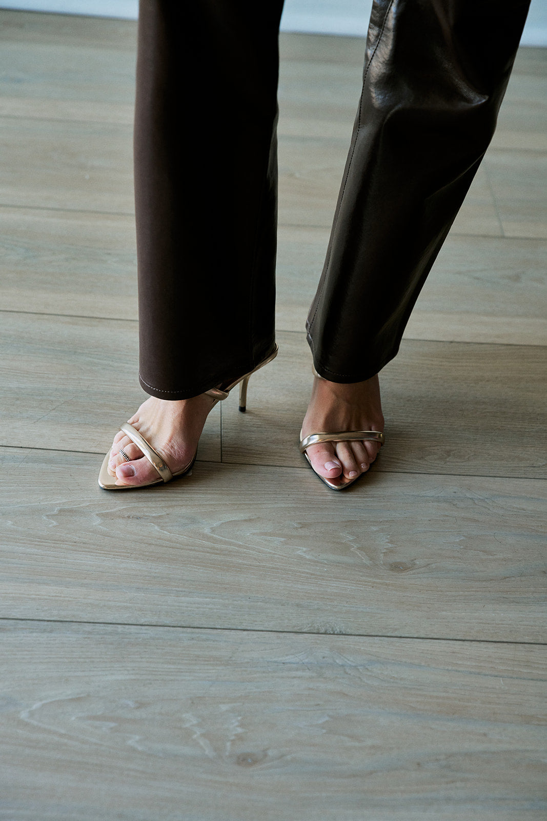 A close-up of a person's feet wearing metallic gold strappy high-heeled sandals and dark brown pants, standing on a light gray wood plank floor.