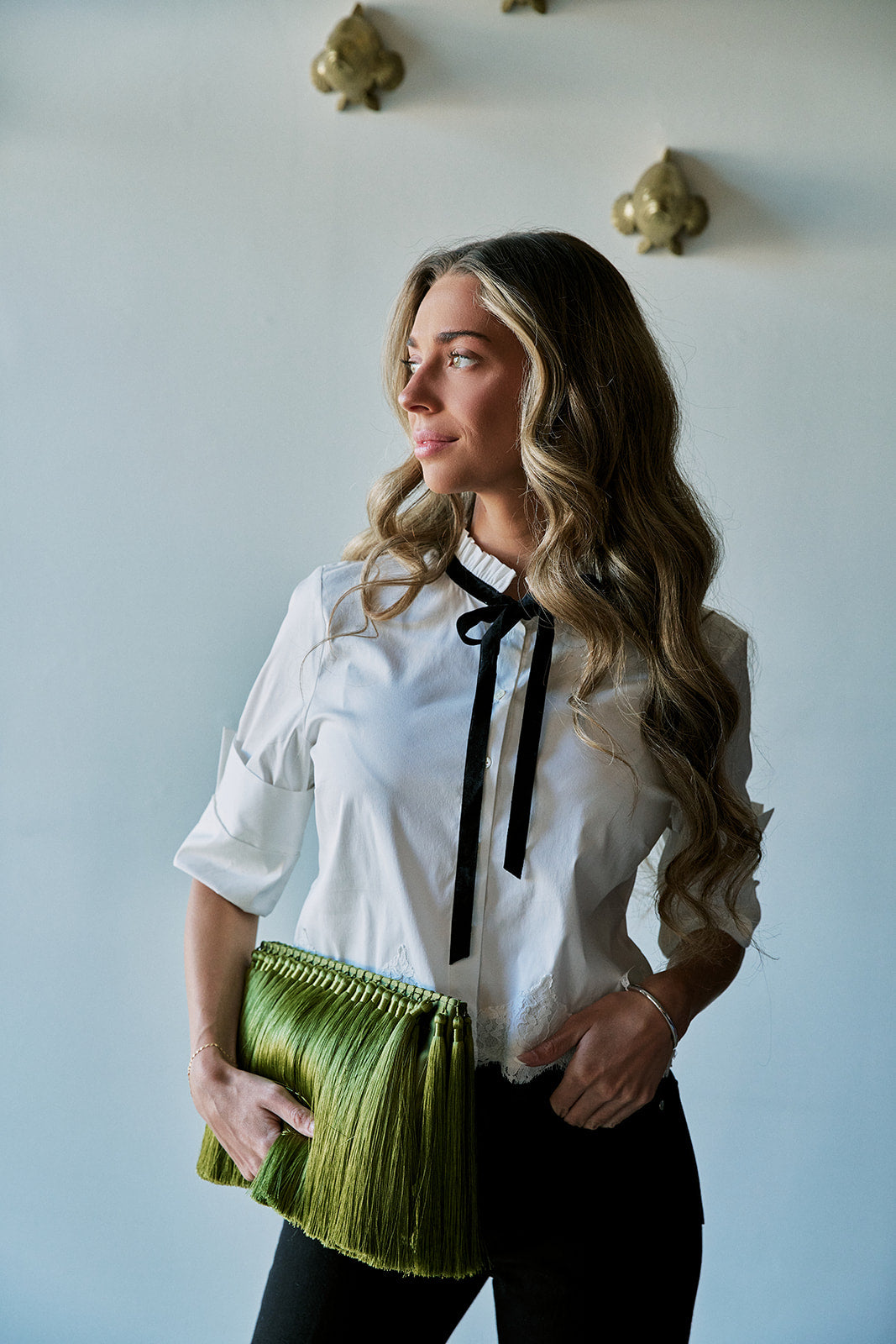 model wearing a white top with a black tie at the neck and black bottoms holding a large green tassel clutch 