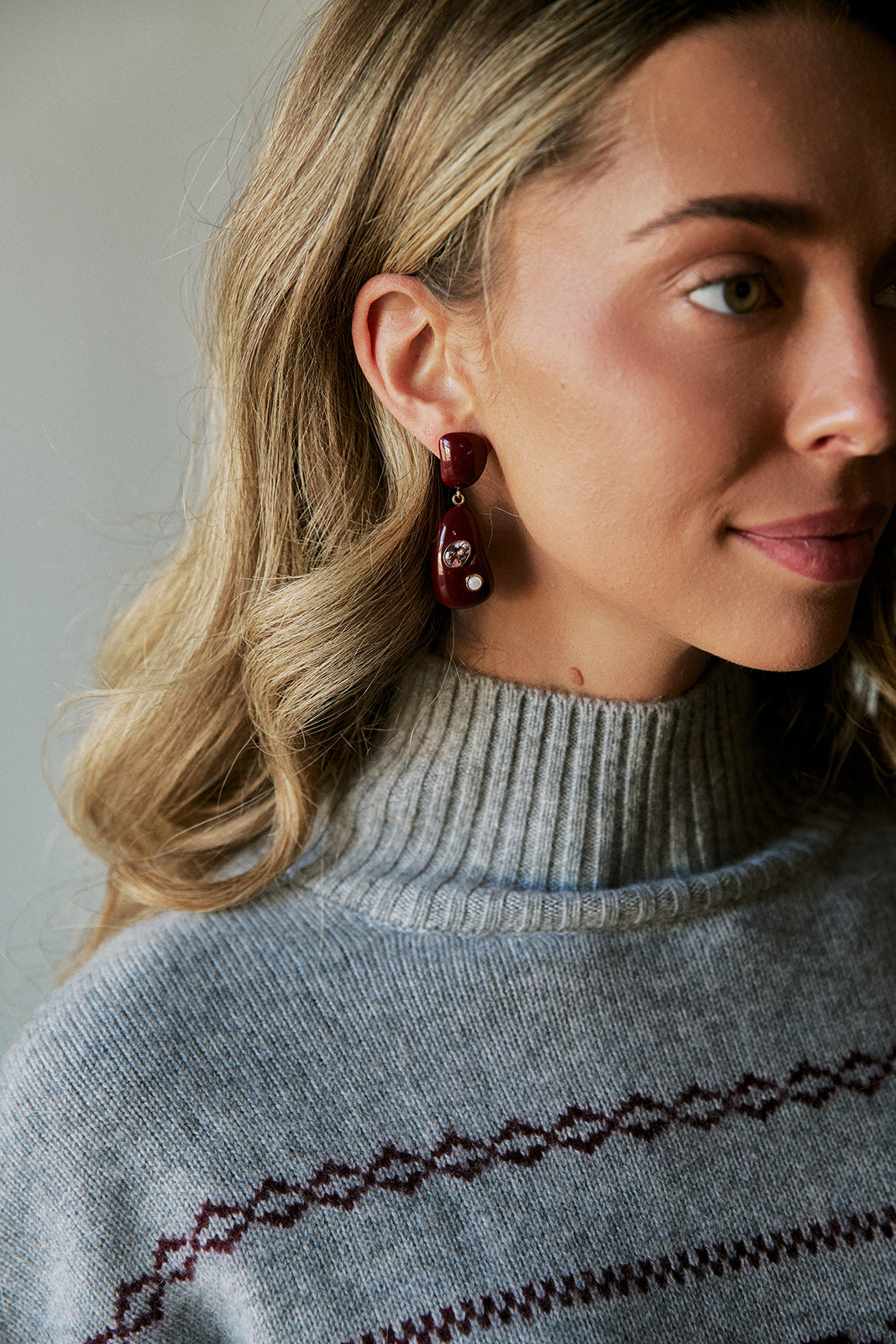 A close-up of a woman wearing a gray knit turtleneck sweater with a burgundy geometric pattern and matching dangling earrings.