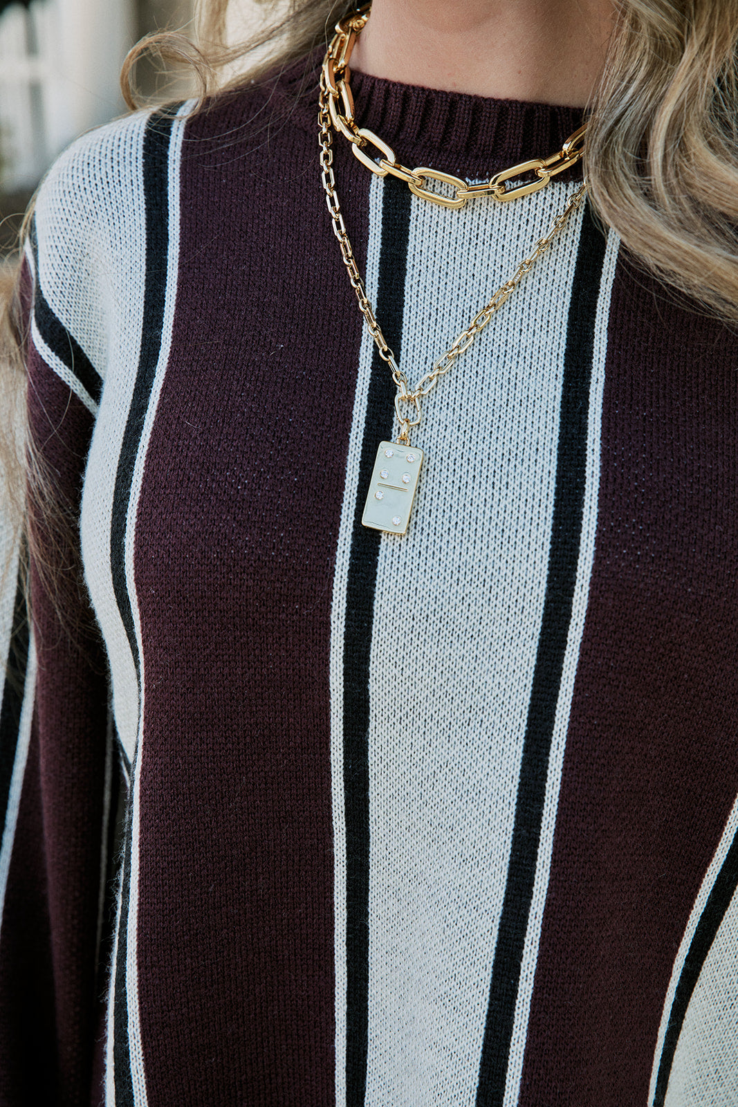 Close-up of a person wearing layered gold necklaces over a burgundy, white, and black striped knit sweater.