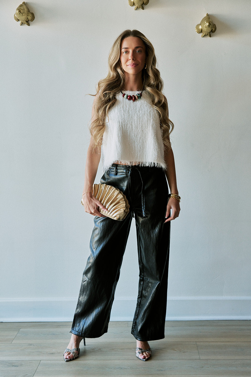 A woman stands indoors wearing a white textured sleeveless top, black wide-leg faux-leather pants, and holding a beige pleated clutch. She is posed against a light gray wall with decorative gold wall pieces and wood flooring visible.