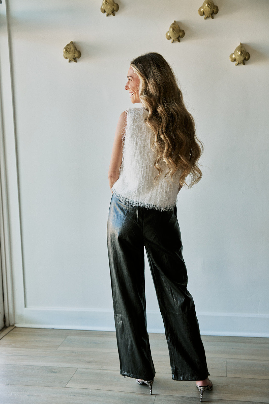 A woman with long wavy brown hair is shown from the back, wearing a sleeveless white textured top and shiny black wide-leg pants, standing in a room with a white wall and light wood floor.