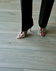 A person wearing black trousers and metallic silver high-heeled sandals with a floral print, standing on a light gray wood plank floor.