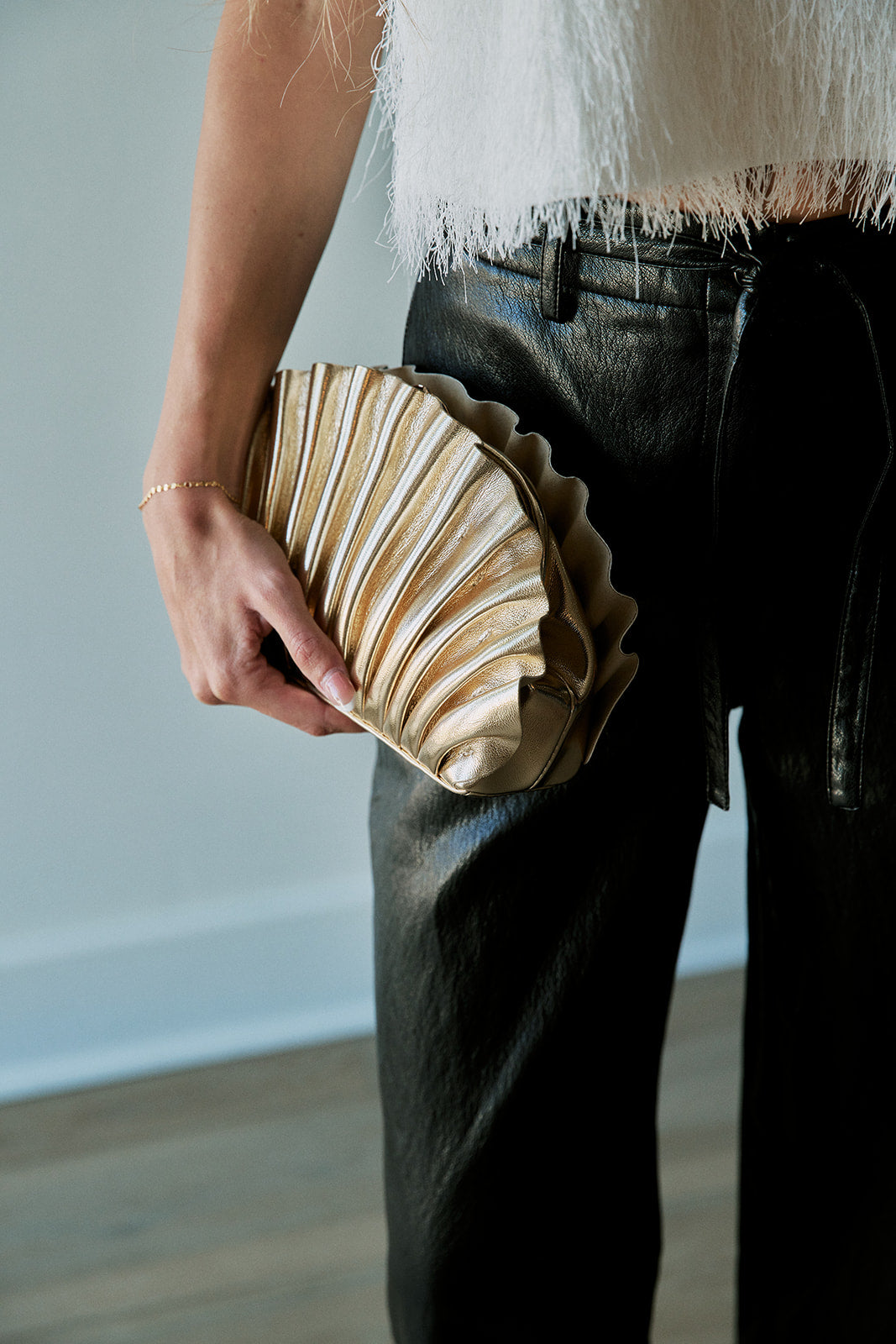 A person holding a pleated, scalloped clutch purse in a light beige or cream tone, paired with black leather pants and a white fringed top.