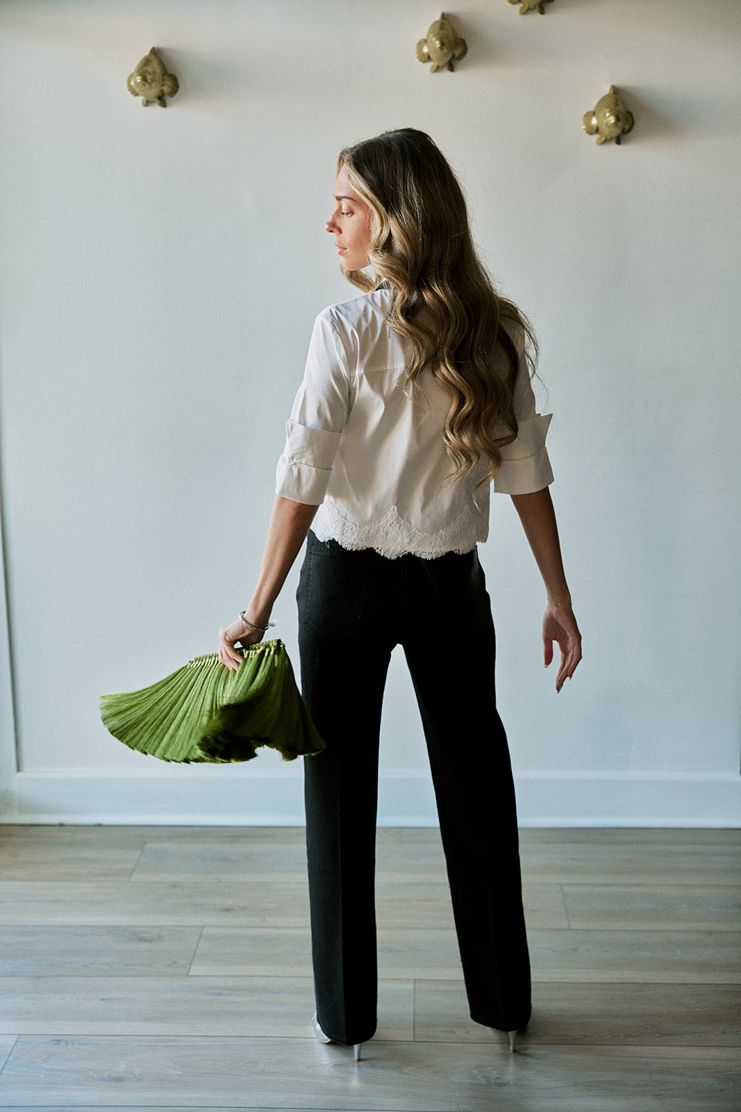 A woman stands in profile wearing a cream ruffled blouse, black wide-leg trousers, and holding a green pleated clutch against a white wall with gold wall decor.