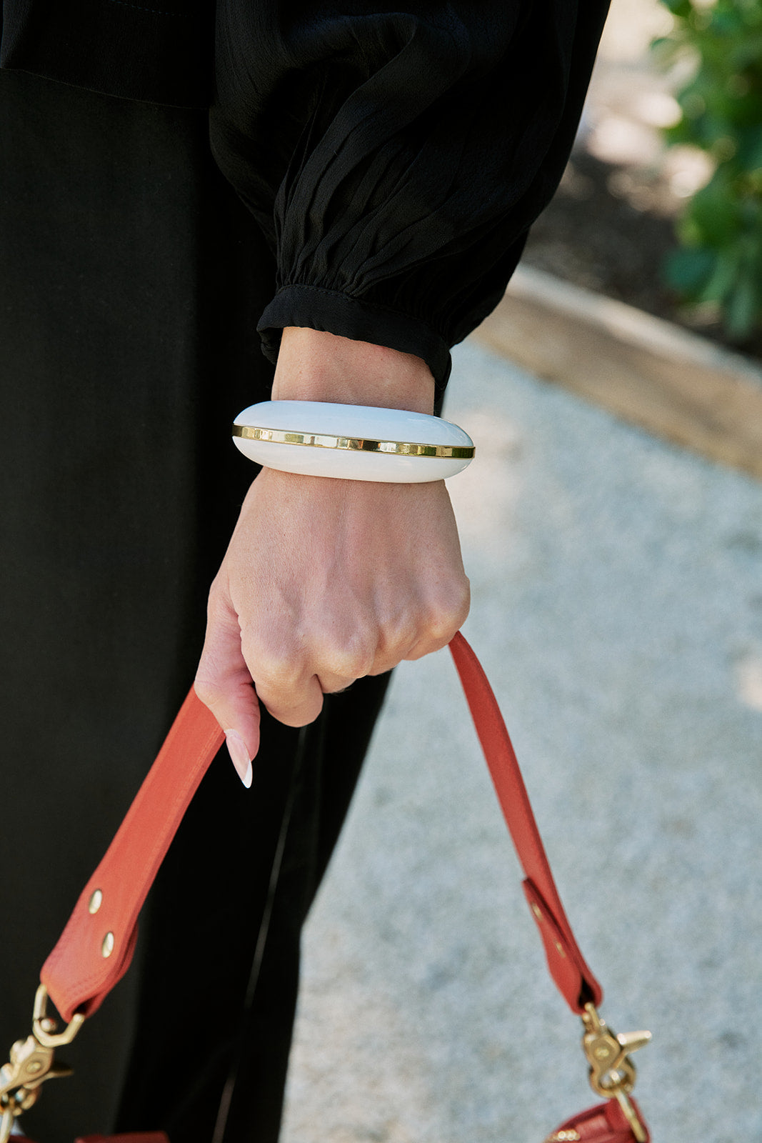 A close-up of a person's hand wearing a chunky white oval bangle with a thin gold band, holding a red leather handbag strap against a blurred outdoor background.