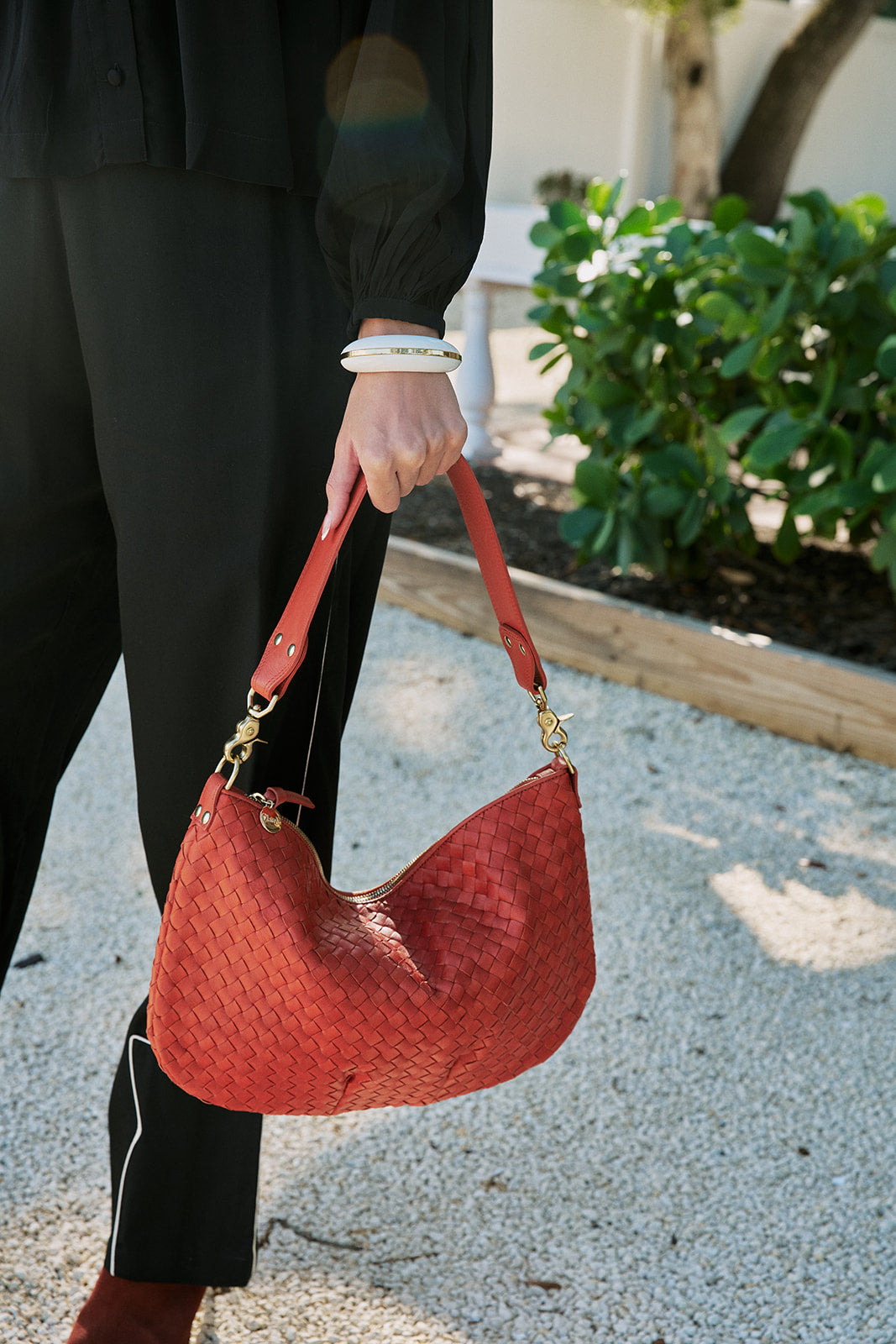 A person in black clothing holds a red woven leather shoulder bag with gold-tone hardware in an outdoor setting with gravel and greenery.