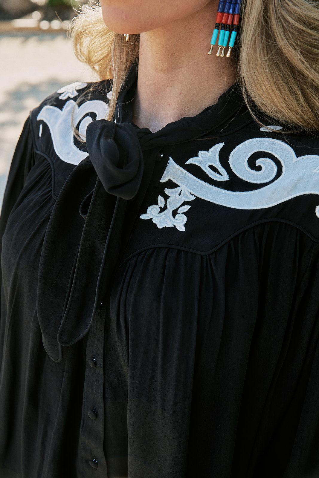 Close-up of a woman wearing a black blouse with a large black bow at the collar and white scrollwork embroidery across the chest. A colorful beaded earring is also visible.