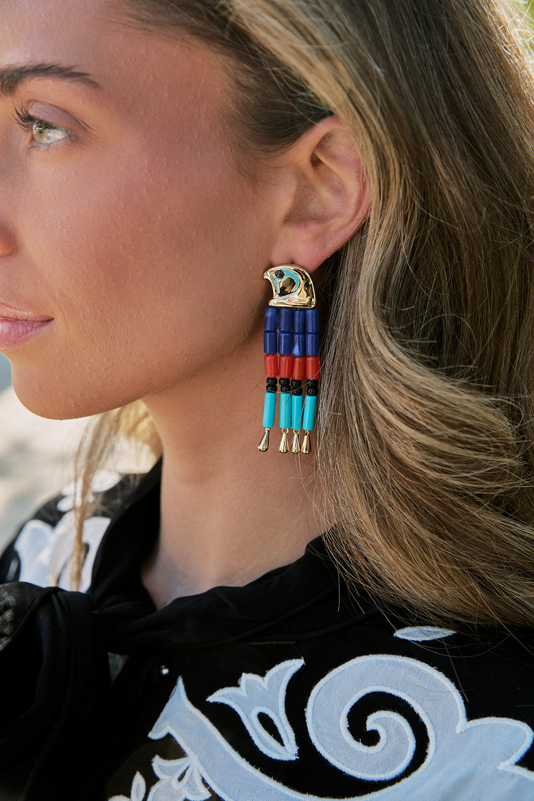 A close-up side profile of a woman wearing a long, dangling statement earring. The earring features a gold-toned eagle head with turquoise and white details, and three vertical strands of beads in blue, red, black, and turquoise, ending in gold-toned tips.