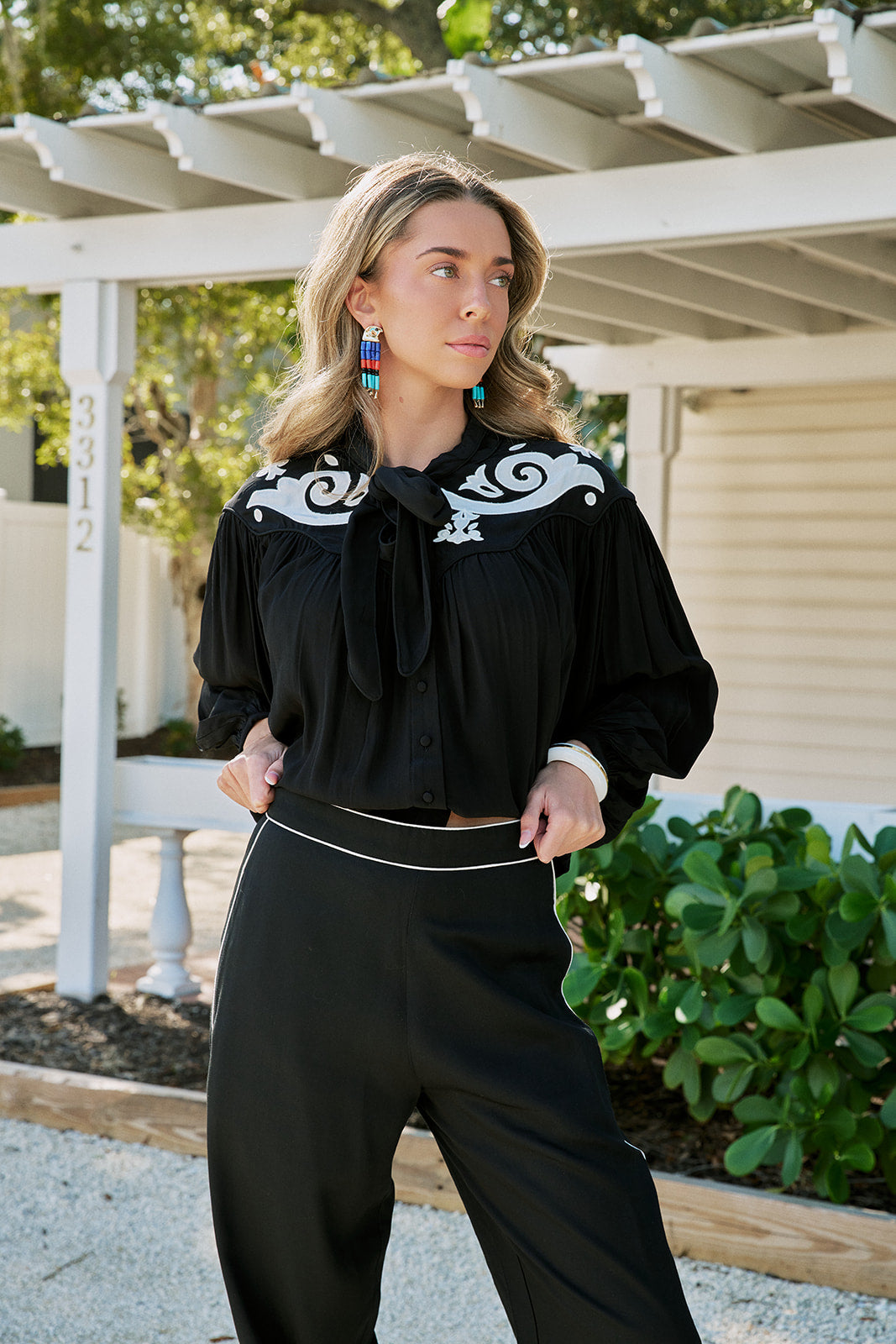 A woman wearing a black long-sleeve blouse with white scrollwork embroidery on the shoulders and a large front bow, paired with black wide-leg pants, standing outdoors under a white pergola.
