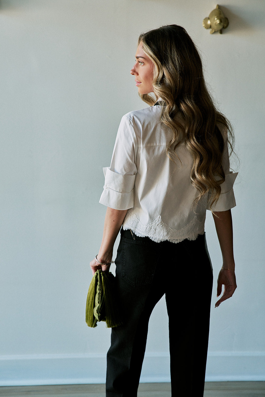 model wearing a white top with a black tie at the neck and black bottoms holding a large green tassel clutch, back view