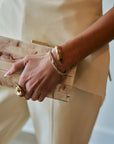 A close-up of a person holding a light wood clutch bag with a gold-tone metal clasp, accessorized with gold bracelets and a ring, wearing a cream-colored dress.