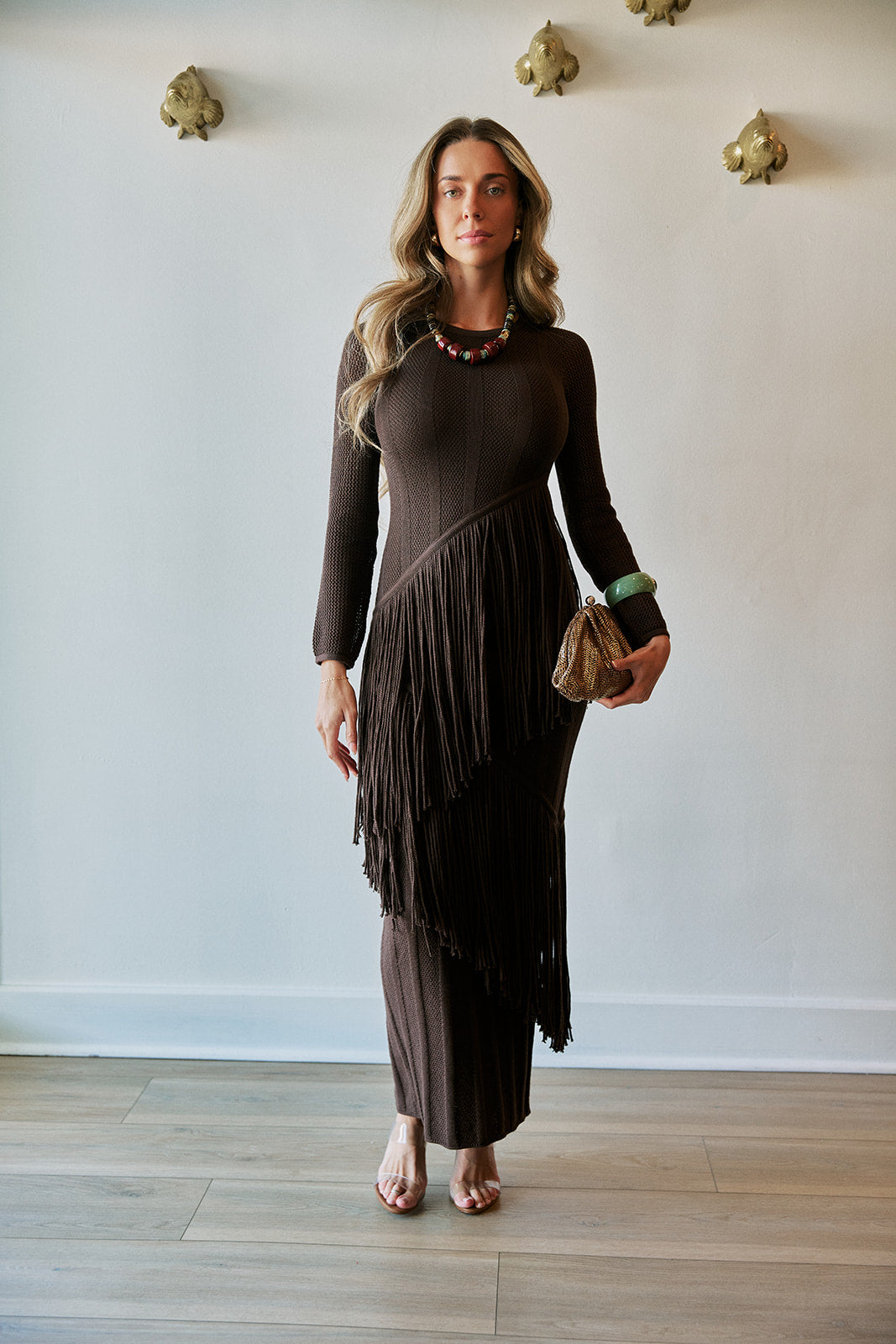 A woman modeling a dark brown long-sleeve knit dress with a layered fringe design, standing in a room with a white wall and light wood floor.