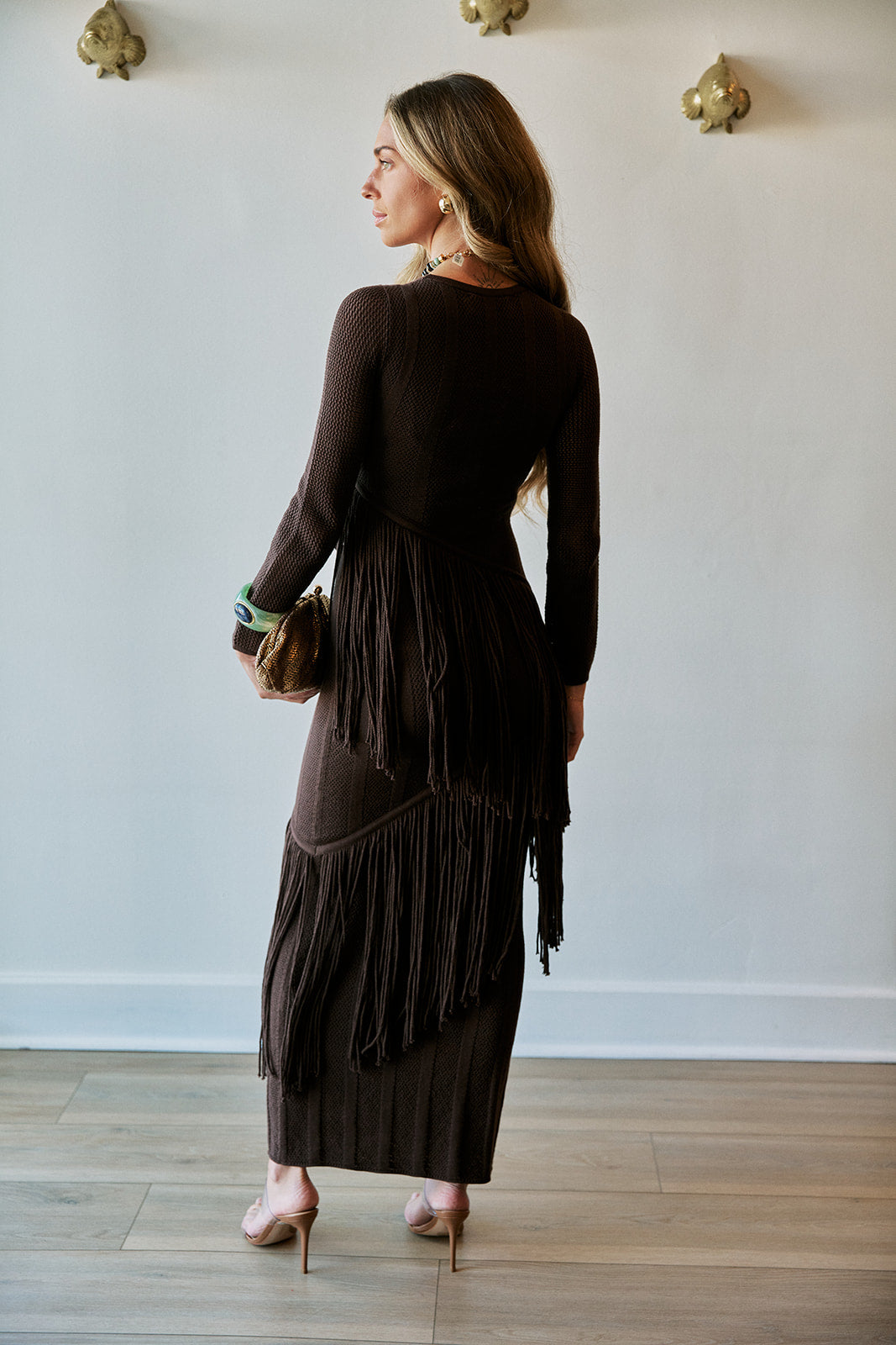 A woman stands with her back to the camera wearing a dark brown long-sleeve maxi dress with fringe detailing along the back and hem. She is holding a small brown clutch and wearing gold high heels in a minimalist room with a white wall and light wood floor.