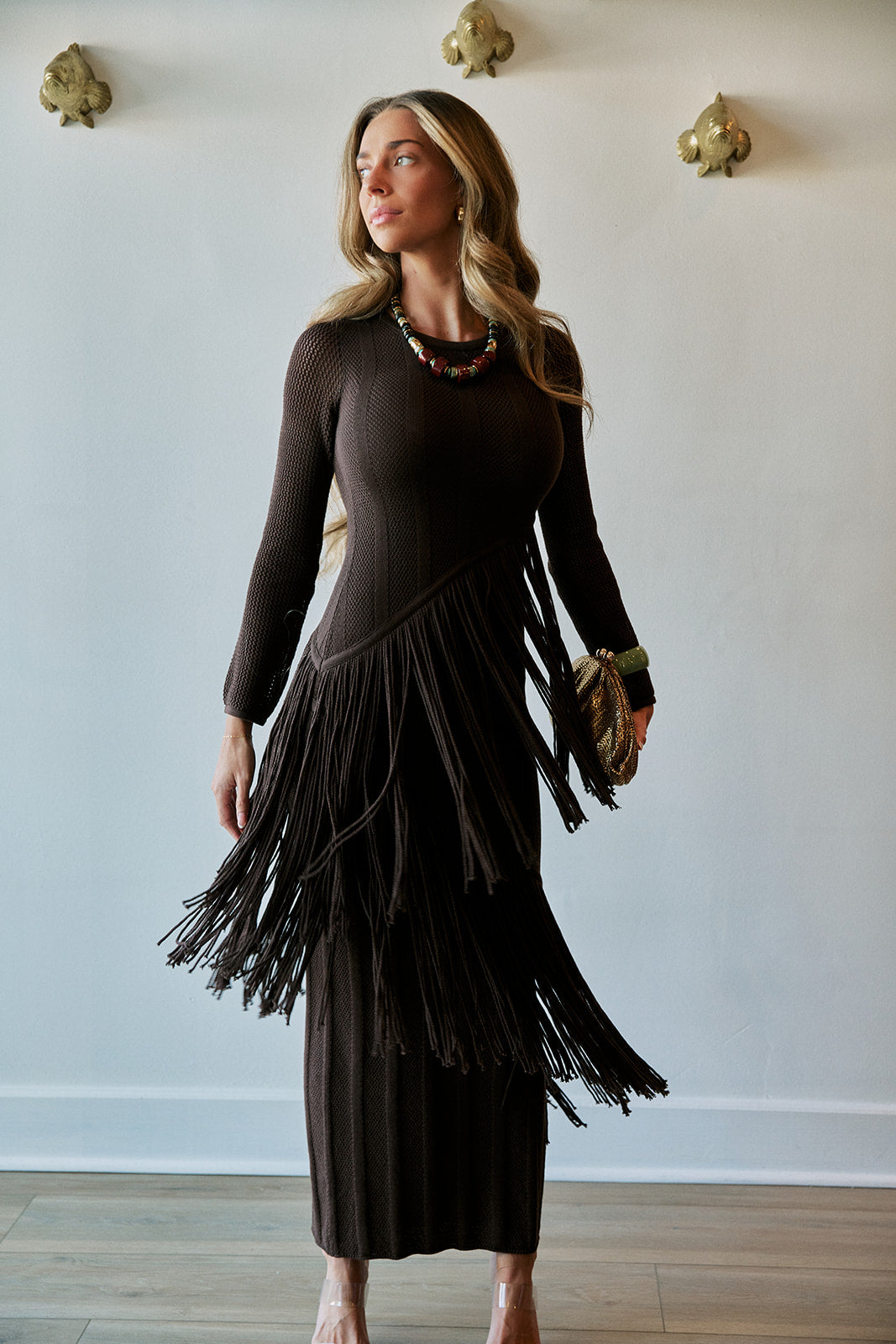 A woman modeling a dark brown long-sleeve midi dress with a large layered fringe skirt, standing against a white wall with gold wall sconces.