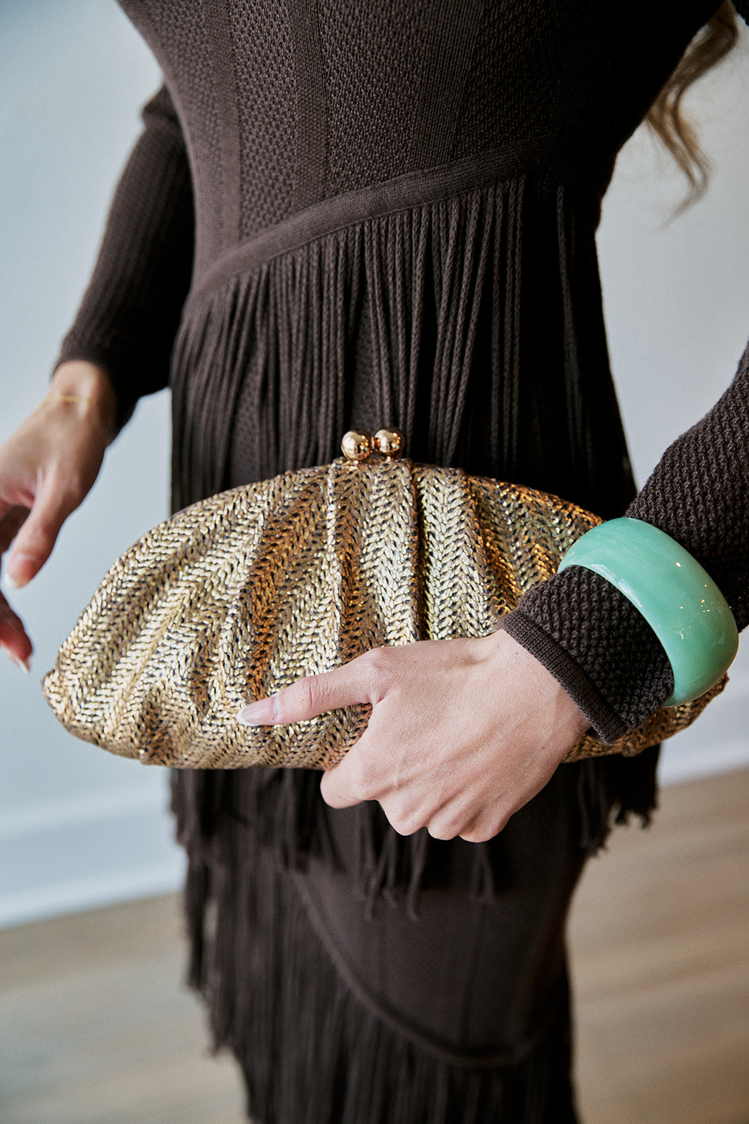 A close-up of a person holding a metallic gold woven clutch purse with a gold-tone clasp. The person is wearing a dark brown fringed knit sweater and a mint green bangle.