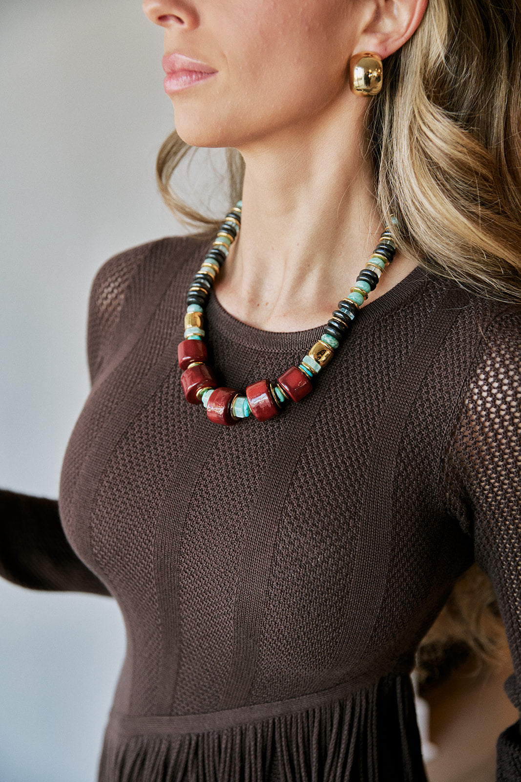 A close-up lifestyle shot of a woman wearing a chunky beaded necklace and matching gold hoop earrings. The necklace features terracotta red, turquoise blue, and dark brown beads, paired with gold-toned spacer beads, worn over a brown knit top.