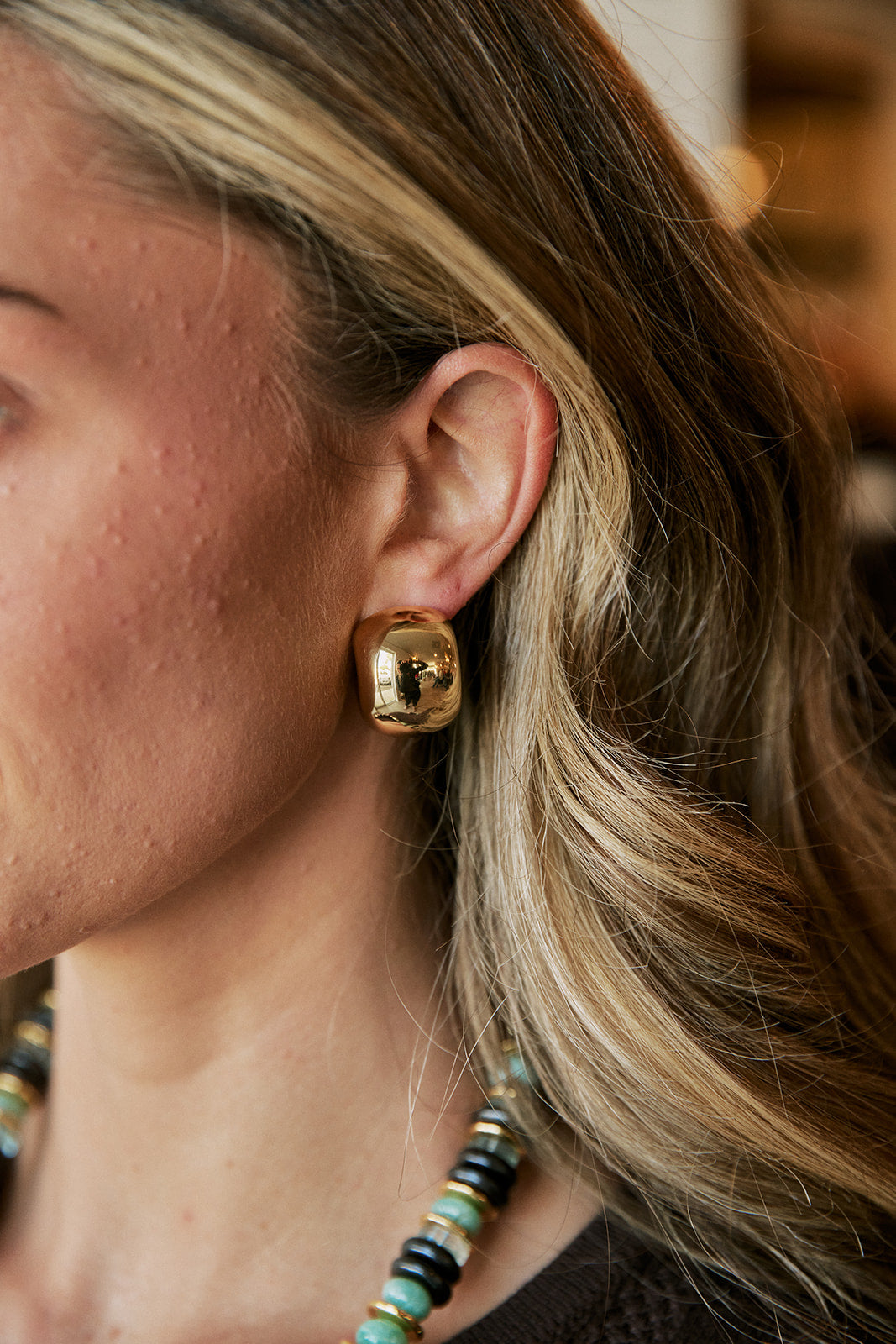 A close-up of a woman's ear wearing a chunky, polished gold dome stud earring, with her blonde hair and a beaded necklace visible.