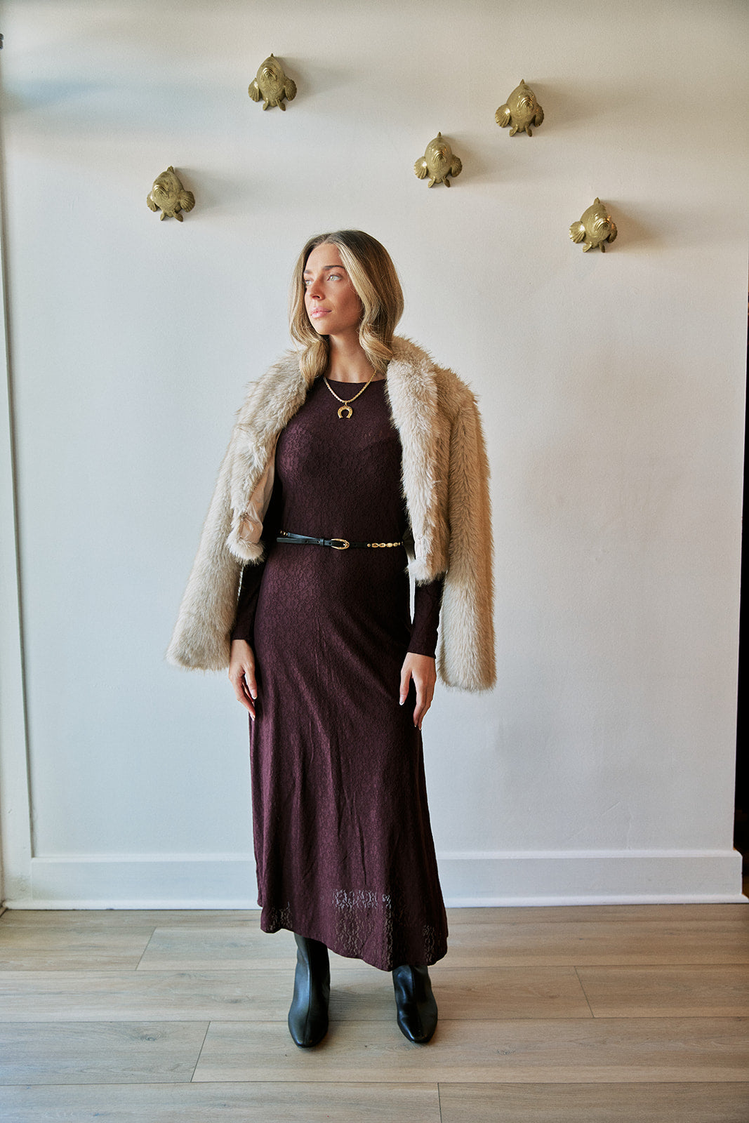 A woman stands indoors wearing a long dark plum dress with a thin black belt, a cream faux-fur jacket, and black ankle boots, posed against a white wall with decorative gold wall mounts.