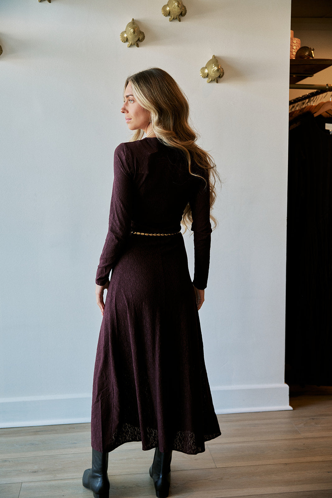 A woman wearing a long-sleeved dark plum midi dress with a gold chain belt, shown from the back in an indoor setting with a white wall and wood floor.