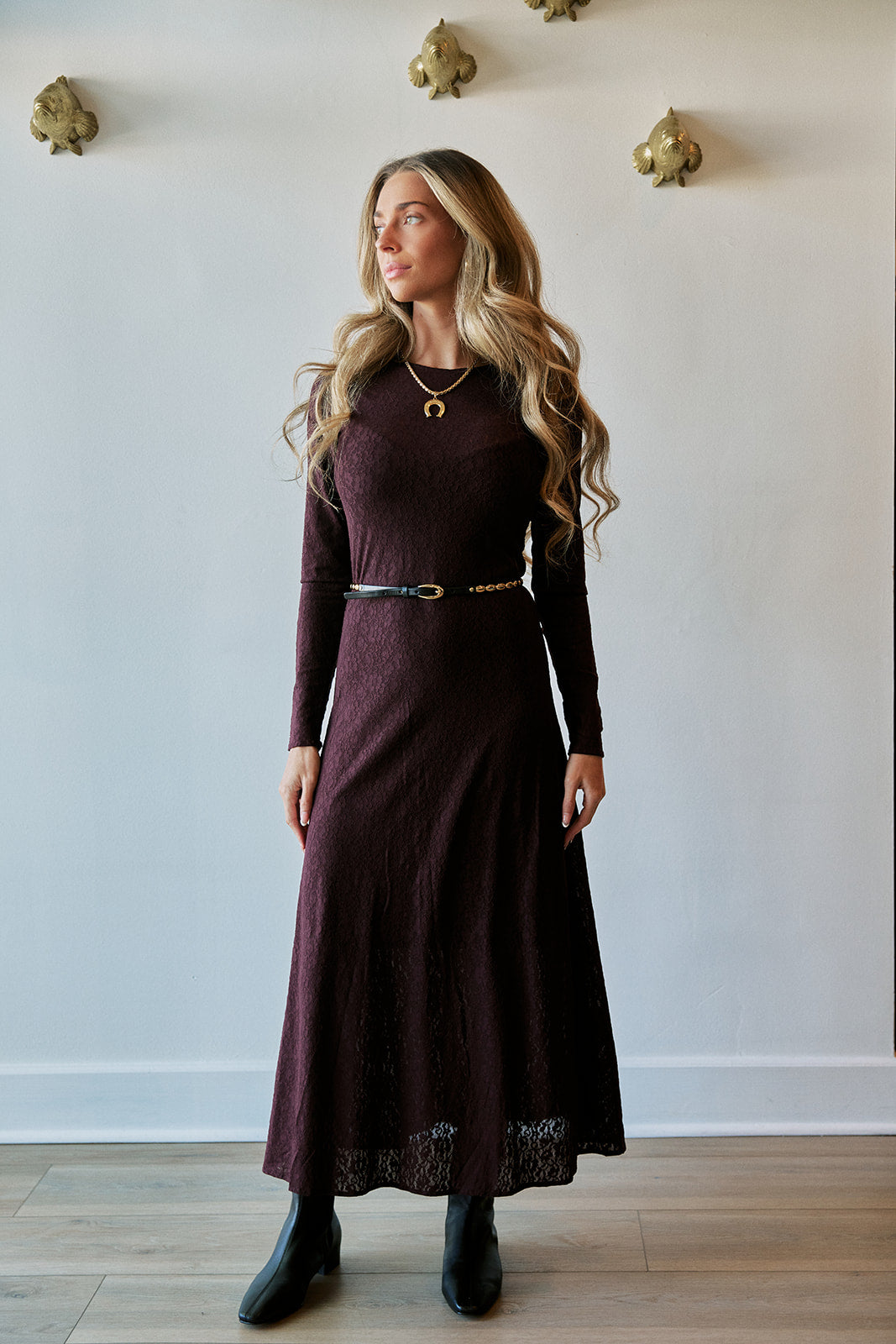 A woman modeling a long-sleeve, high-neck, floor-length burgundy dress with a thin belt at the waist and black ankle boots, standing against a white wall with gold decorative accents.