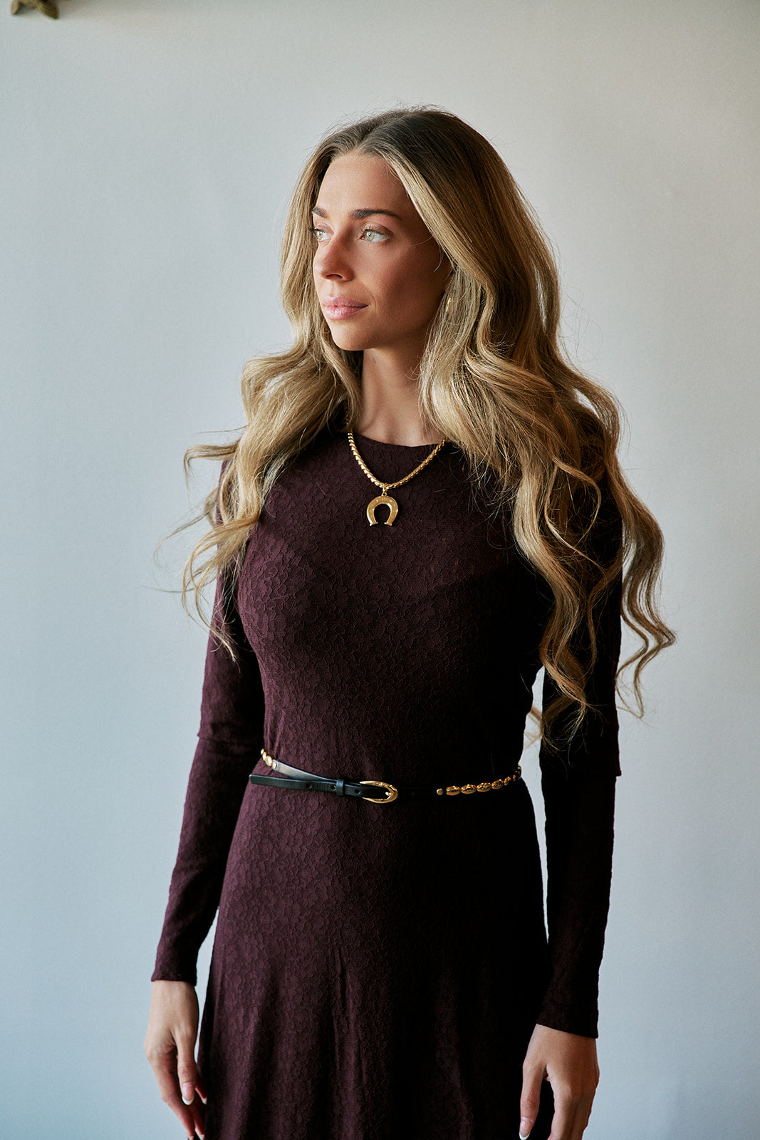 A woman with long wavy blonde hair wearing a dark burgundy long-sleeve textured dress with a thin belt, standing against a plain light gray wall.