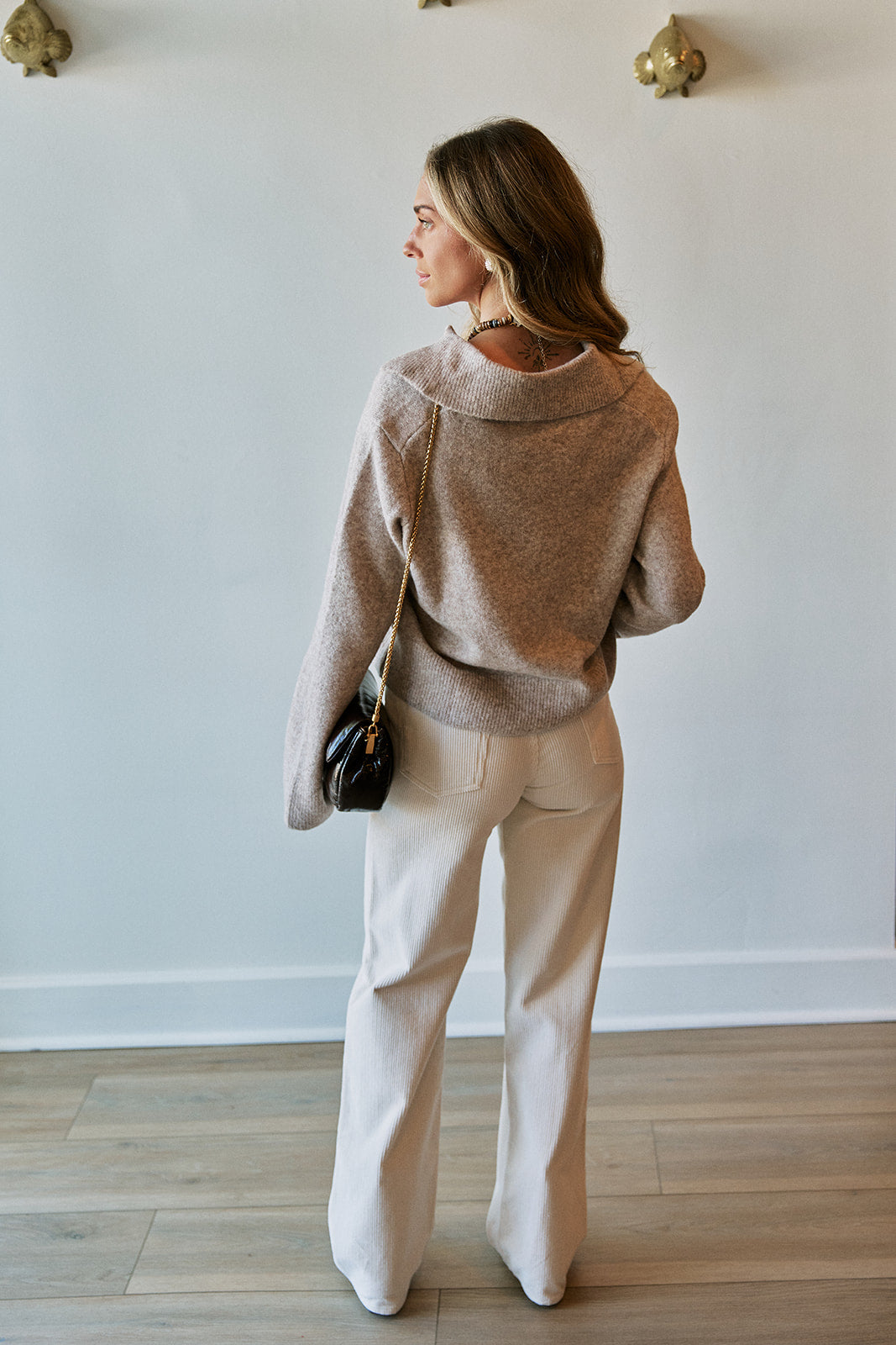 A woman stands with her back to the camera wearing a fuzzy taupe knit sweater and cream wide-leg trousers, holding a small black shoulder bag in a bright room with light wood floors.