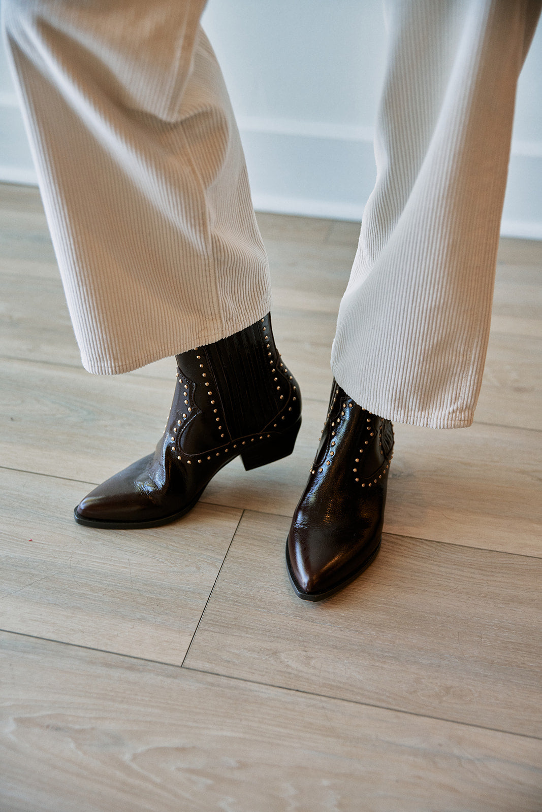A person wearing dark cherry brown pointed-toe ankle boots with sstud detailing and white ribbed wide-leg pants, standing on a light wood floor.