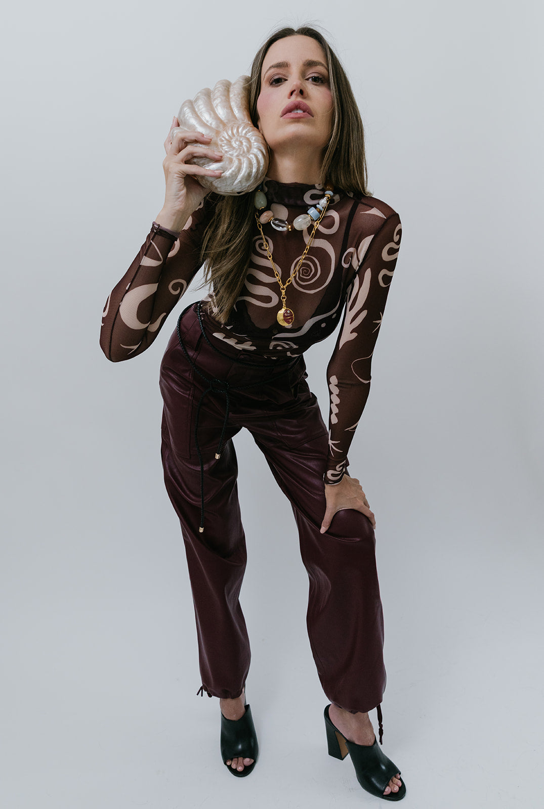 Woman holding a shell clutch wearing maroon pants and patterned top against a plain background