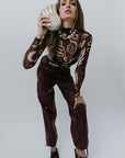 Woman holding a shell clutch wearing maroon pants and patterned top against a plain background