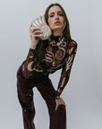Woman in a patterned top and brown pants holding a shell clutch against a plain background
