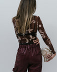 Person wearing a patterned sheer top and maroon pants holding a clutch against a white background back view