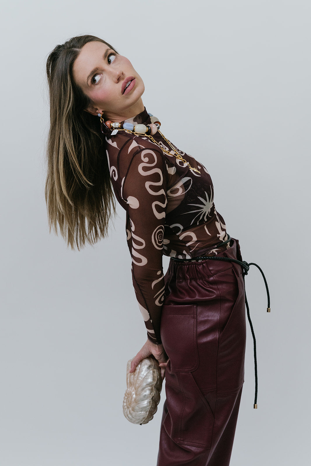 Woman wearing a patterned sheer top and maroon pants against a plain background side view