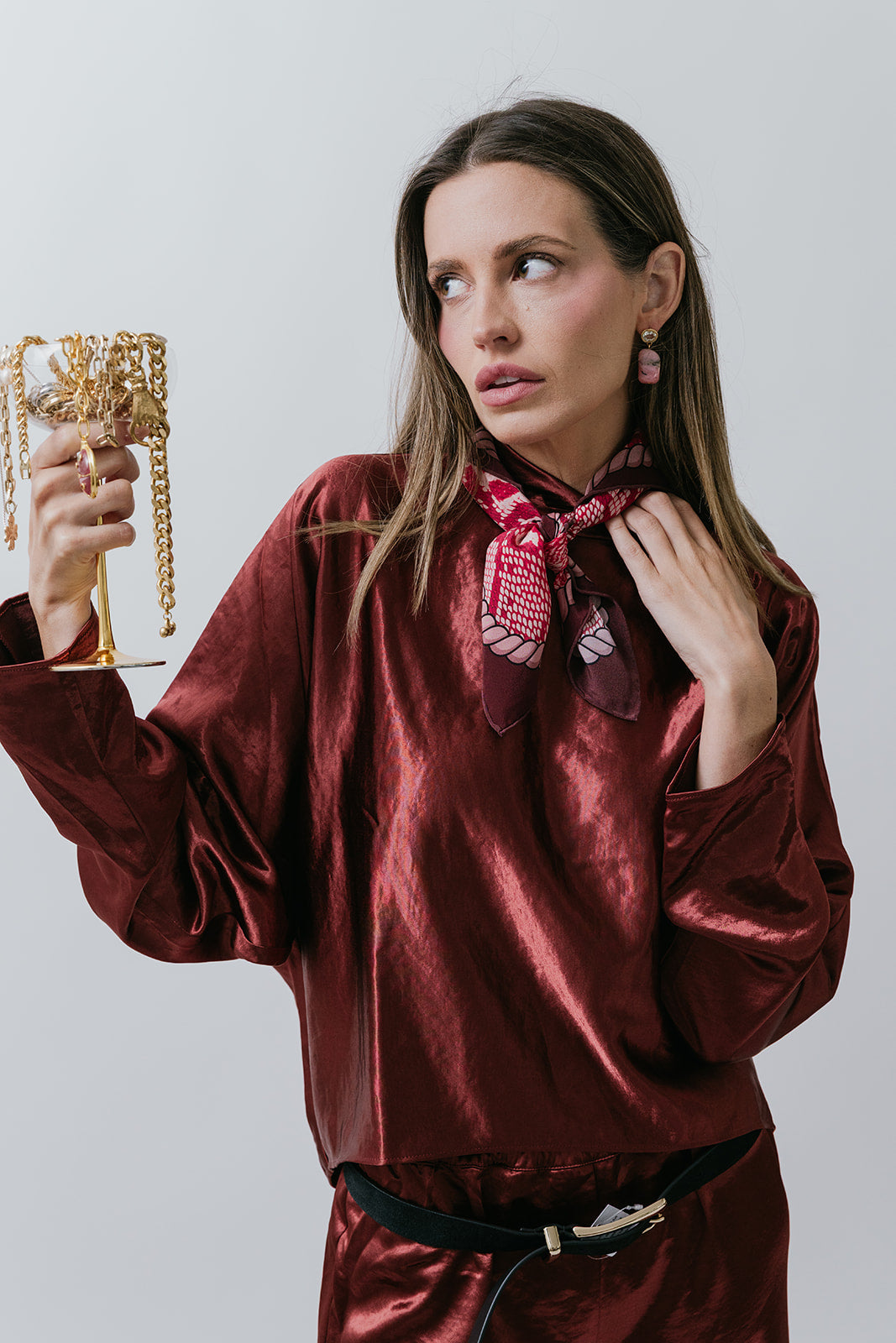 Woman in a burgundy satin outfit and neck scarf holding necklaces with a white background