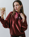 Woman in a burgundy satin outfit and neck scarf holding necklaces with a white background