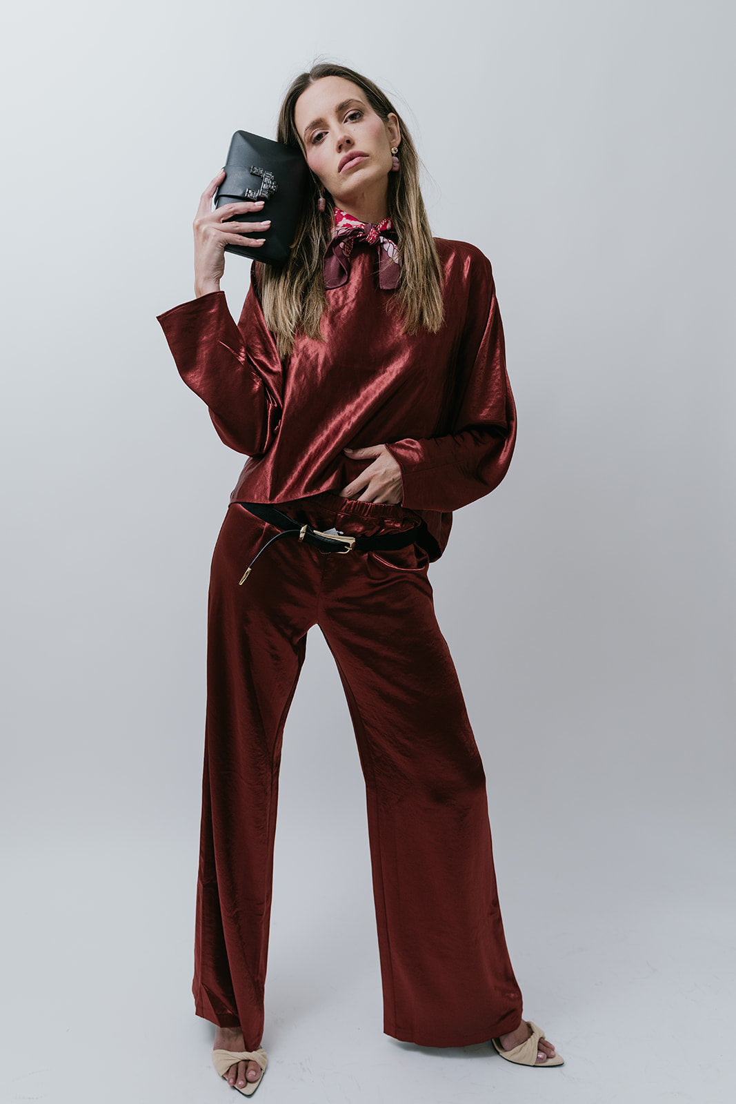 Woman in a burgundy satin outfit and black clutch posing against a white background