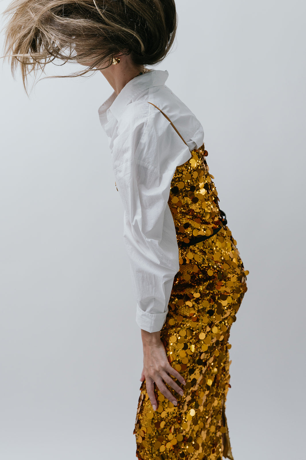 Person wearing a white shirt and gold sequin dress against a plain background side view