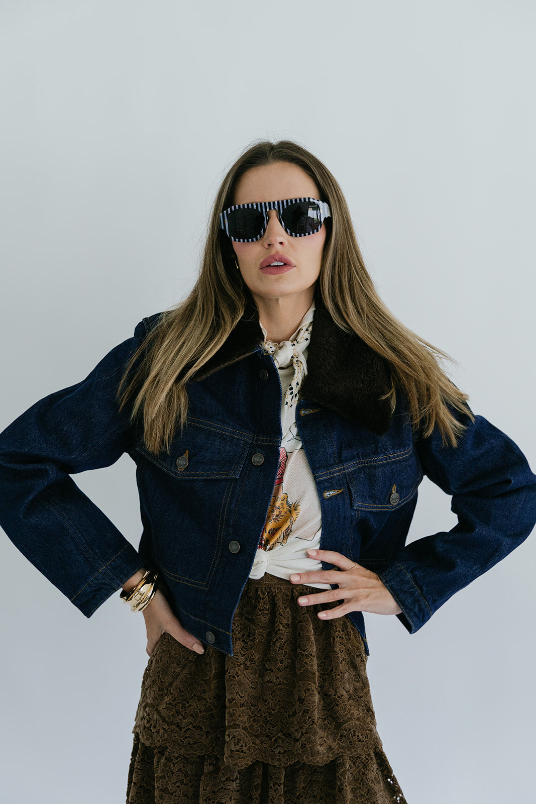 Woman wearing sunglasses, a denim jacket, and a patterned shirt against a white background